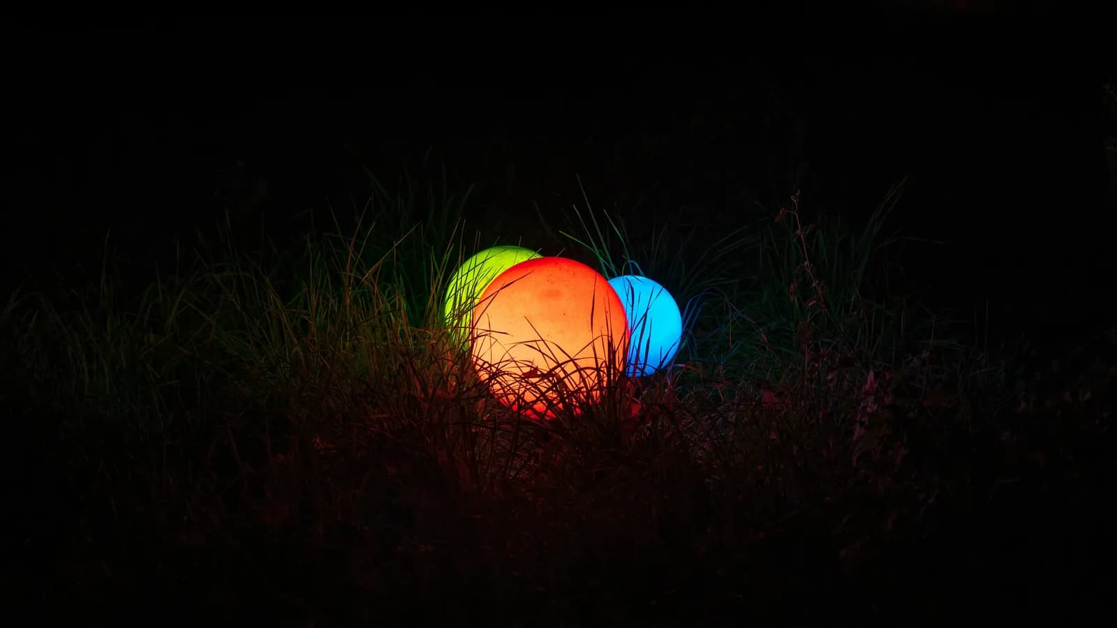 Trois balles lumineuses rouge, verte et bleue posées dans l'herbe en pleine nuit