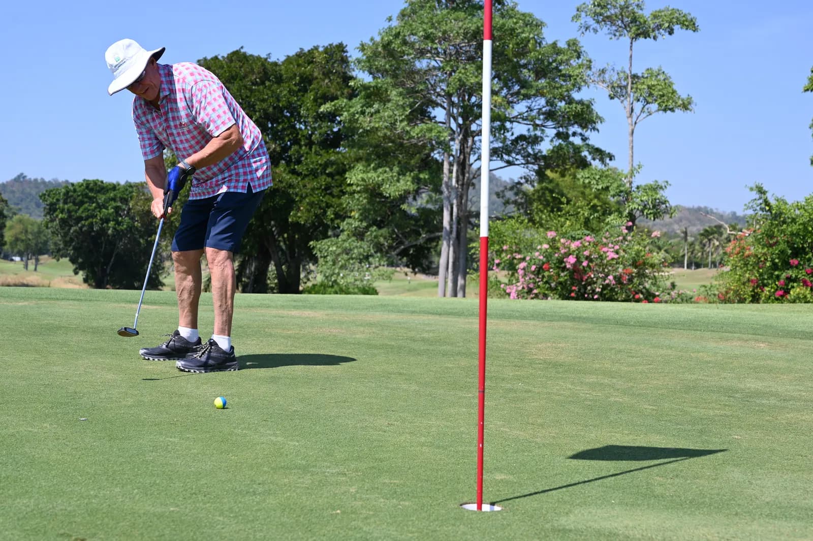 Golfeur aux cheveux gris, chapeau blanc et chemise à carreaux roses, qui putte sur un green ensoleillé à côté du drapeau rouge et blanc