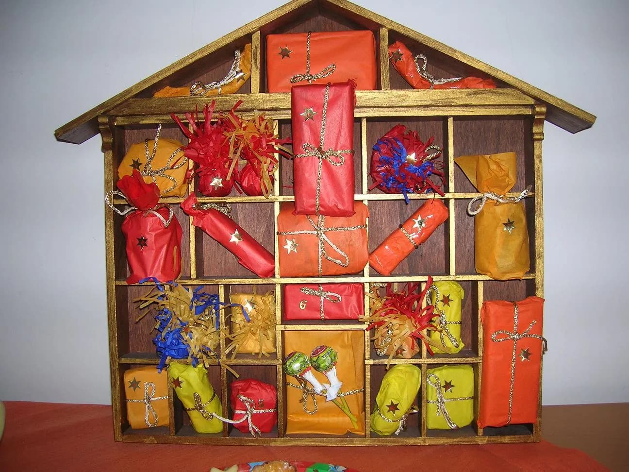 Calendrier de l'Avent fait maison en forme de maison en bois, dont les petites cases contiennent des paquets emballés de papier rouge, orange et jaune noués d'un fil doré