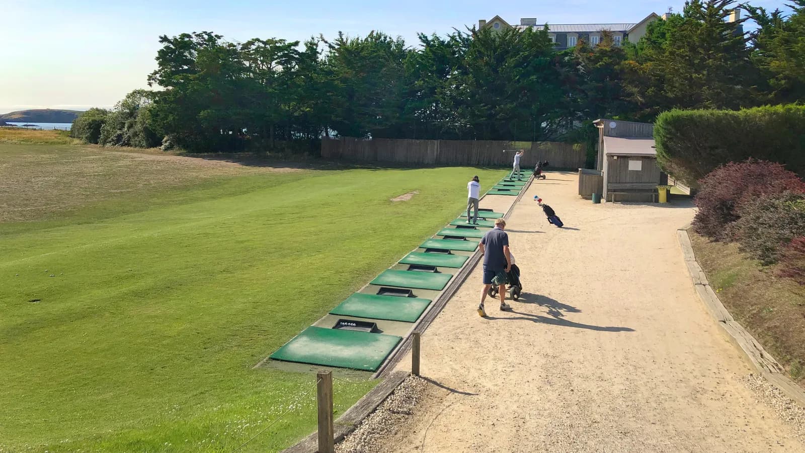 Practice d'un golf du bord de mer en Bretagne : une rangée de tapis de frappe verts le long d'une allée sablée, quelques golfeurs de dos avec leur chariot, et la mer au loin