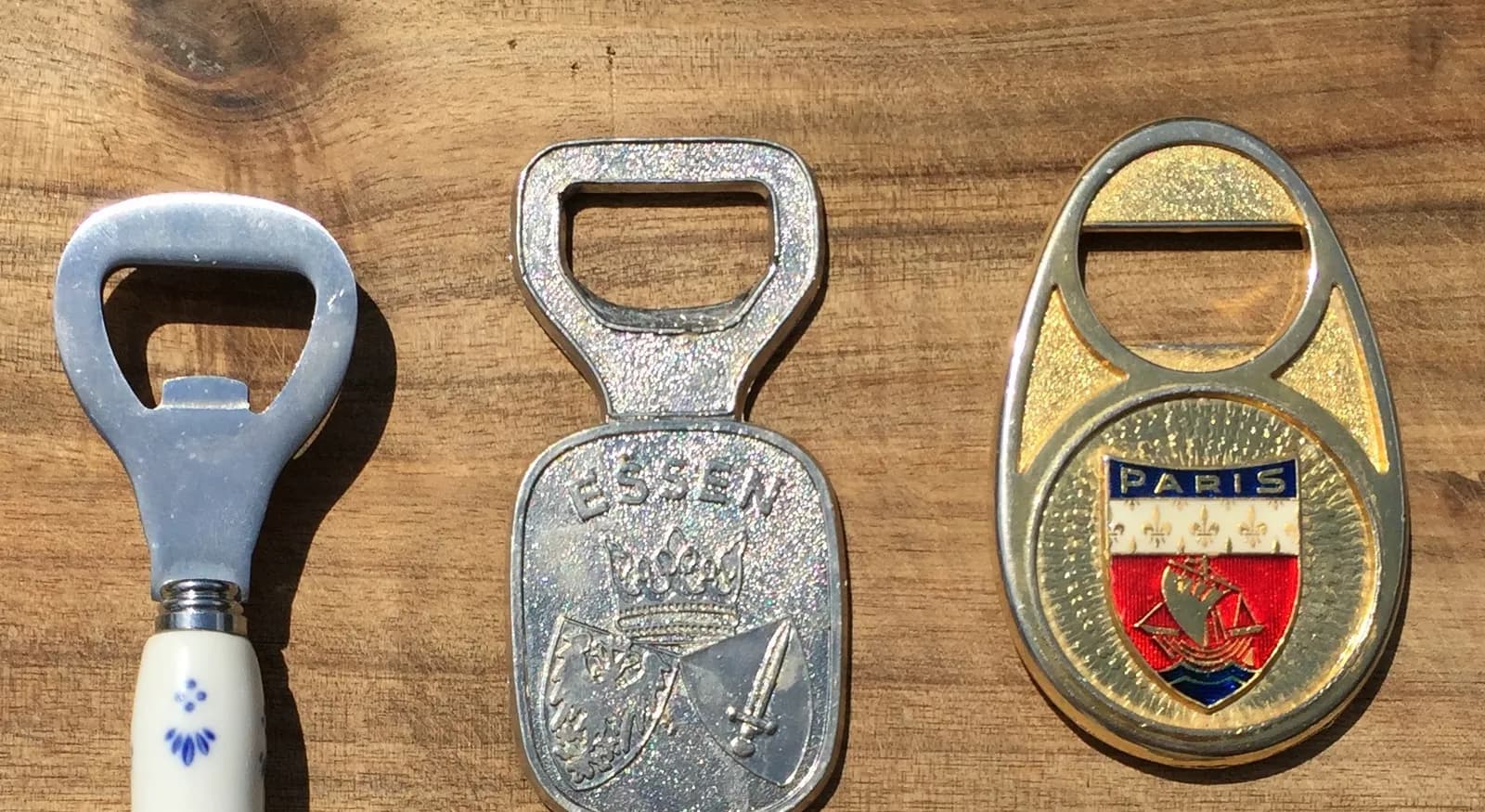 Trois décapsuleurs souvenirs posés sur une planche en bois : un manche en faïence bleu et blanc, un modèle en métal gravé d'un blason et un modèle doré aux armes de Paris