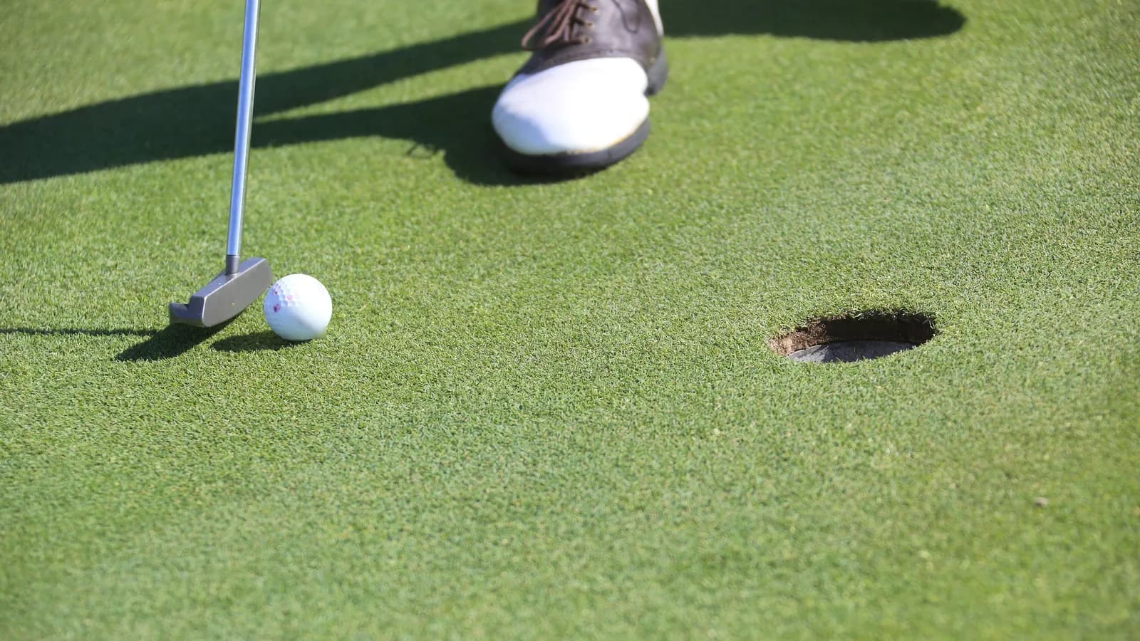 Putter posé derrière une balle blanche sur un green, à un mètre environ du trou, avec les chaussures du golfeur en haut de l'image