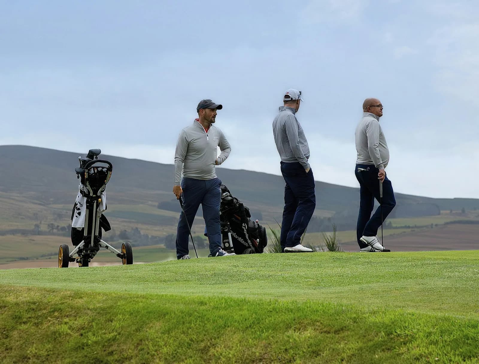 Trois golfeurs debout sur un parcours vallonné à côté de leurs chariots