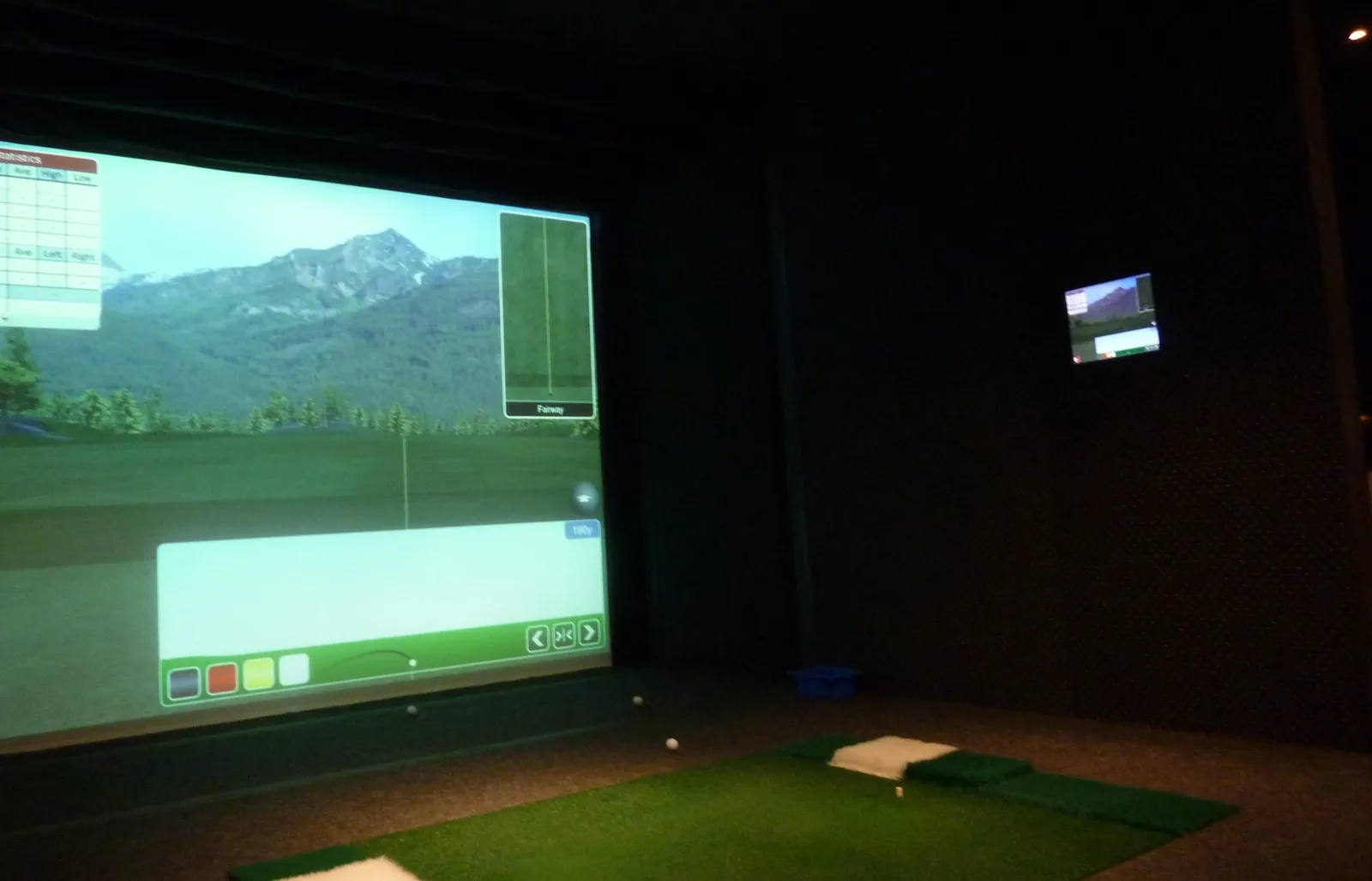 Salle de golf indoor plongée dans la pénombre : un grand écran projette un fairway au pied d'une montagne, devant un tapis de frappe vert avec une balle sur un tee