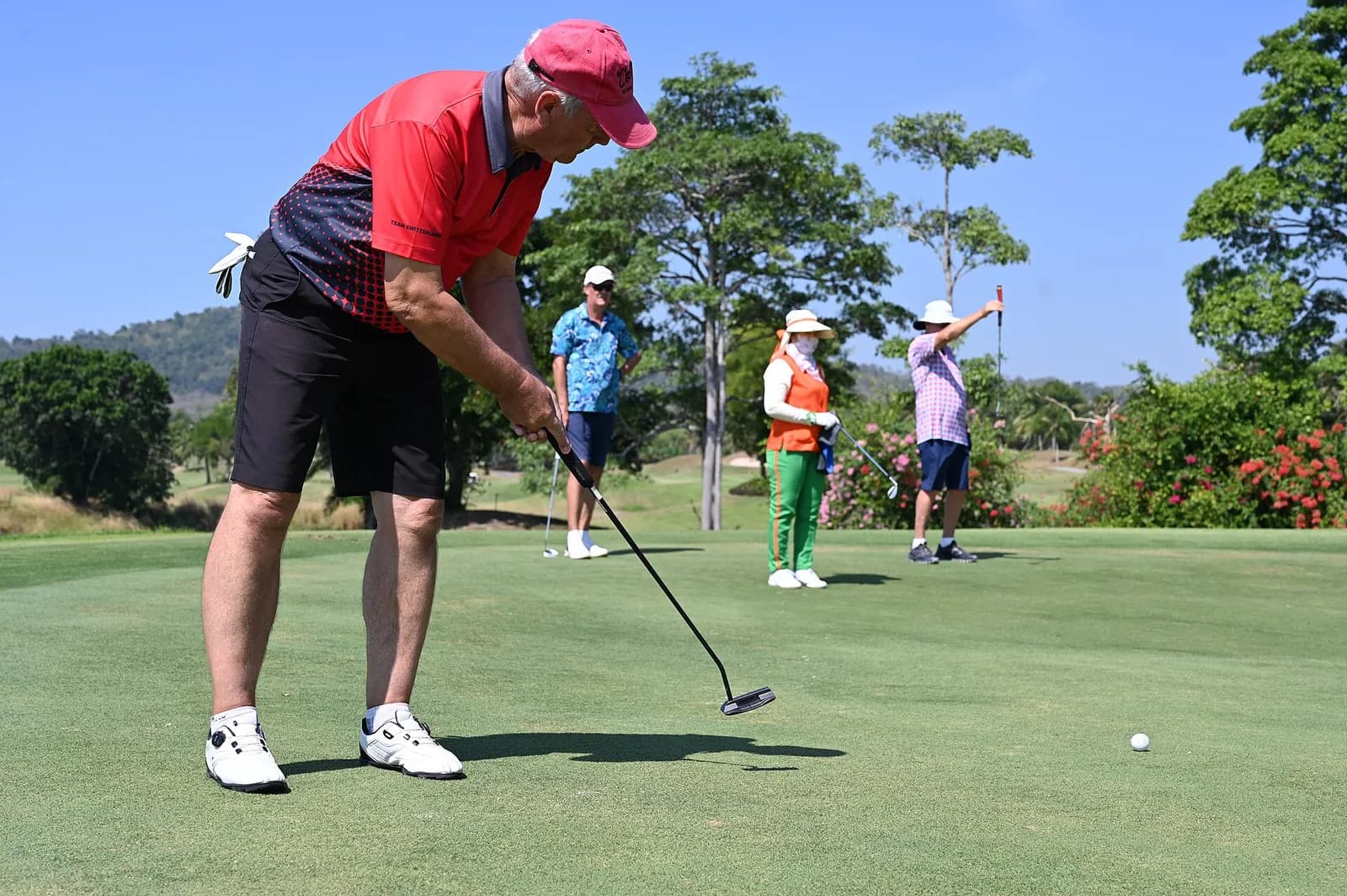 Golfeur en polo rouge qui putte sur un green ensoleillé pendant que trois partenaires de jeu attendent en arrière-plan