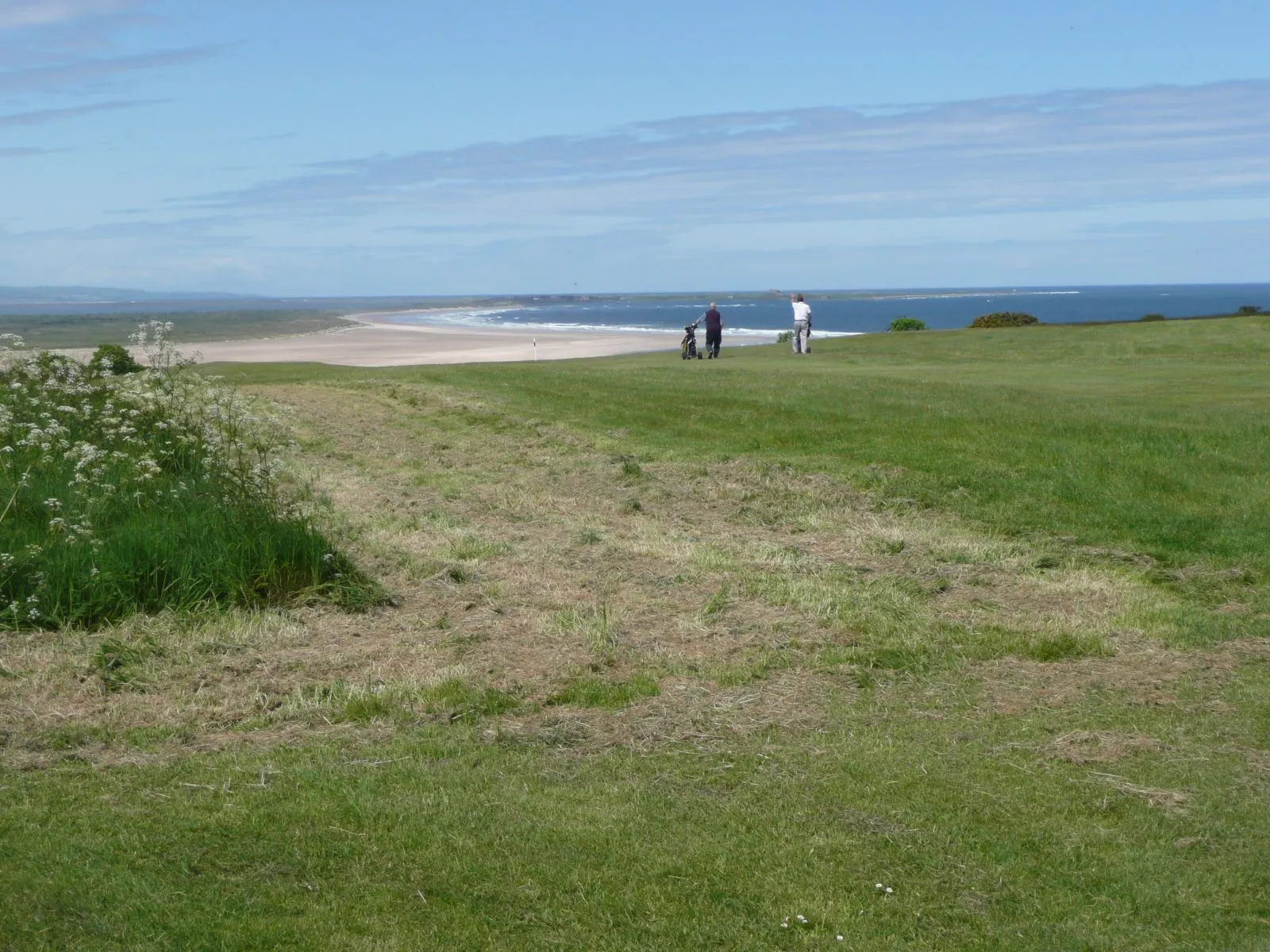 Deux golfeurs, l'un tirant son chariot, marchent sur un fairway en bord de mer, face à une longue plage et à l'océan