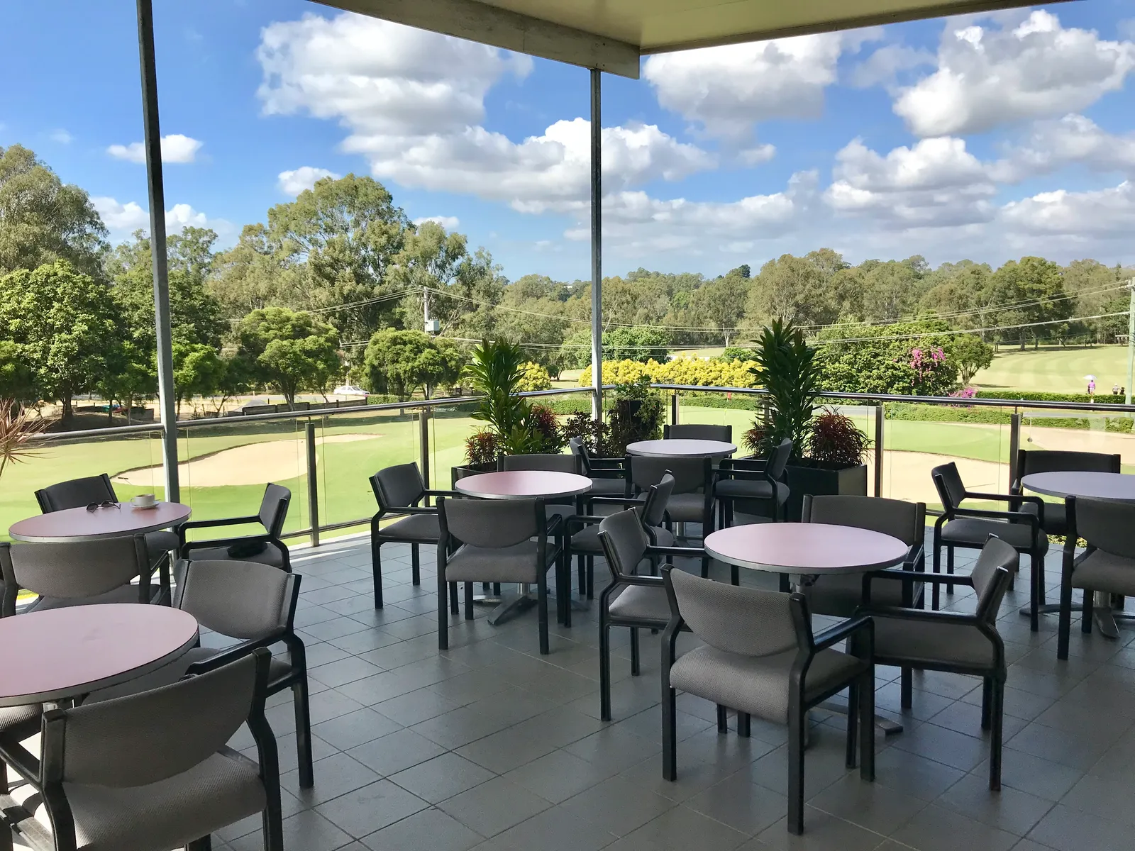 Terrasse couverte d'un club-house de golf avec des tables rondes et des fauteuils gris, ouverte sur un parcours bordé d'arbres et un bunker de sable