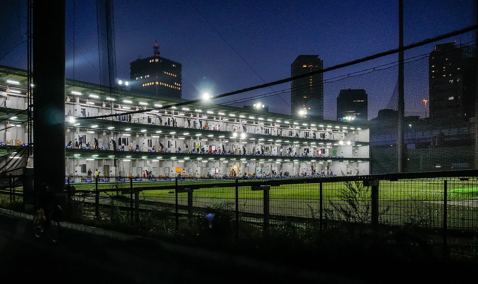 Practice de golf sur trois étages éclairé de nuit, avec des joueurs aux postes de frappe et le terrain illuminé au premier plan