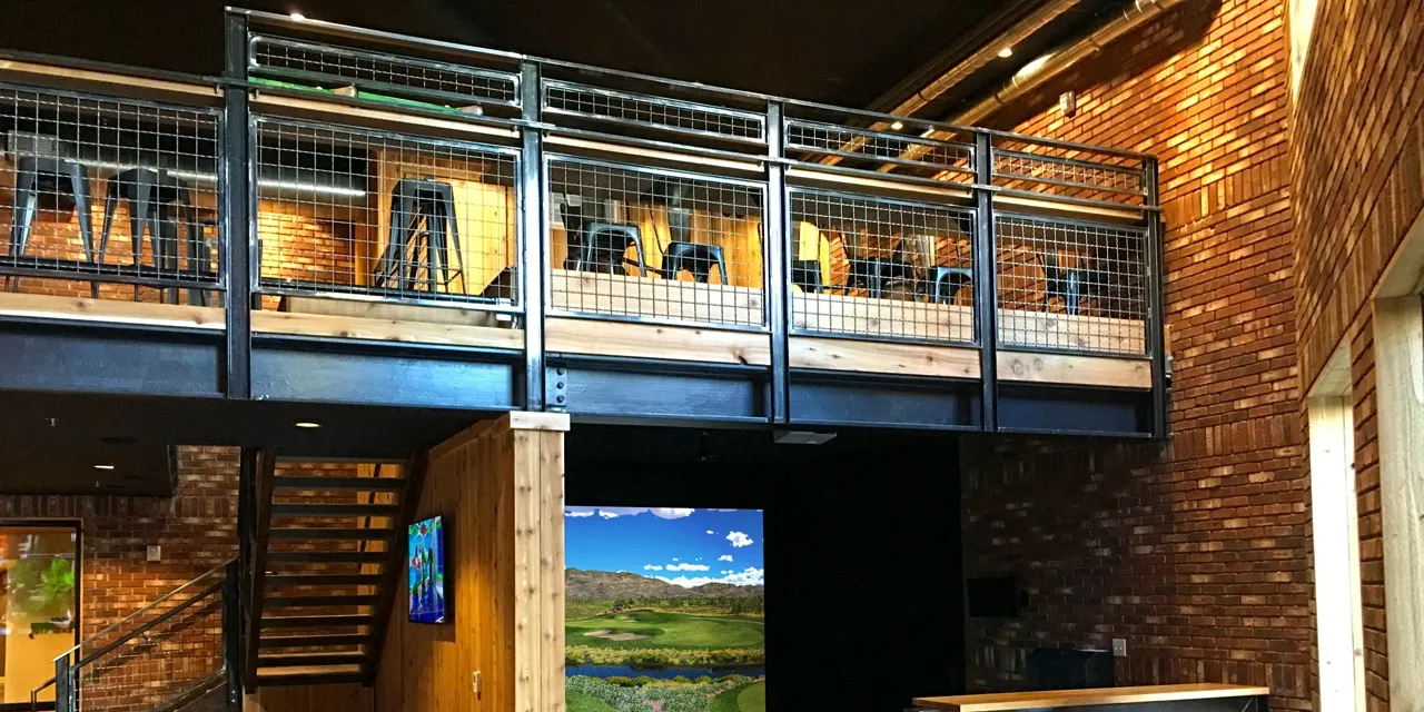 Salle de golf indoor aménagée dans un bâtiment en brique, avec une mezzanine à garde-corps grillagé et un écran de simulateur qui affiche un green entouré d'eau