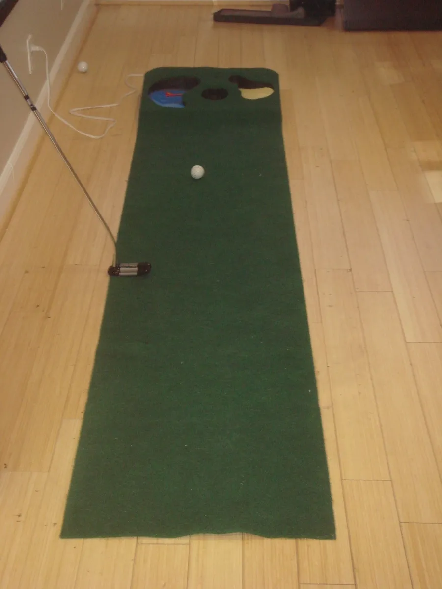 Tapis de putting vert déroulé sur un parquet clair, avec une balle au milieu, un putter posé sur le bord et des trous à l'extrémité du tapis
