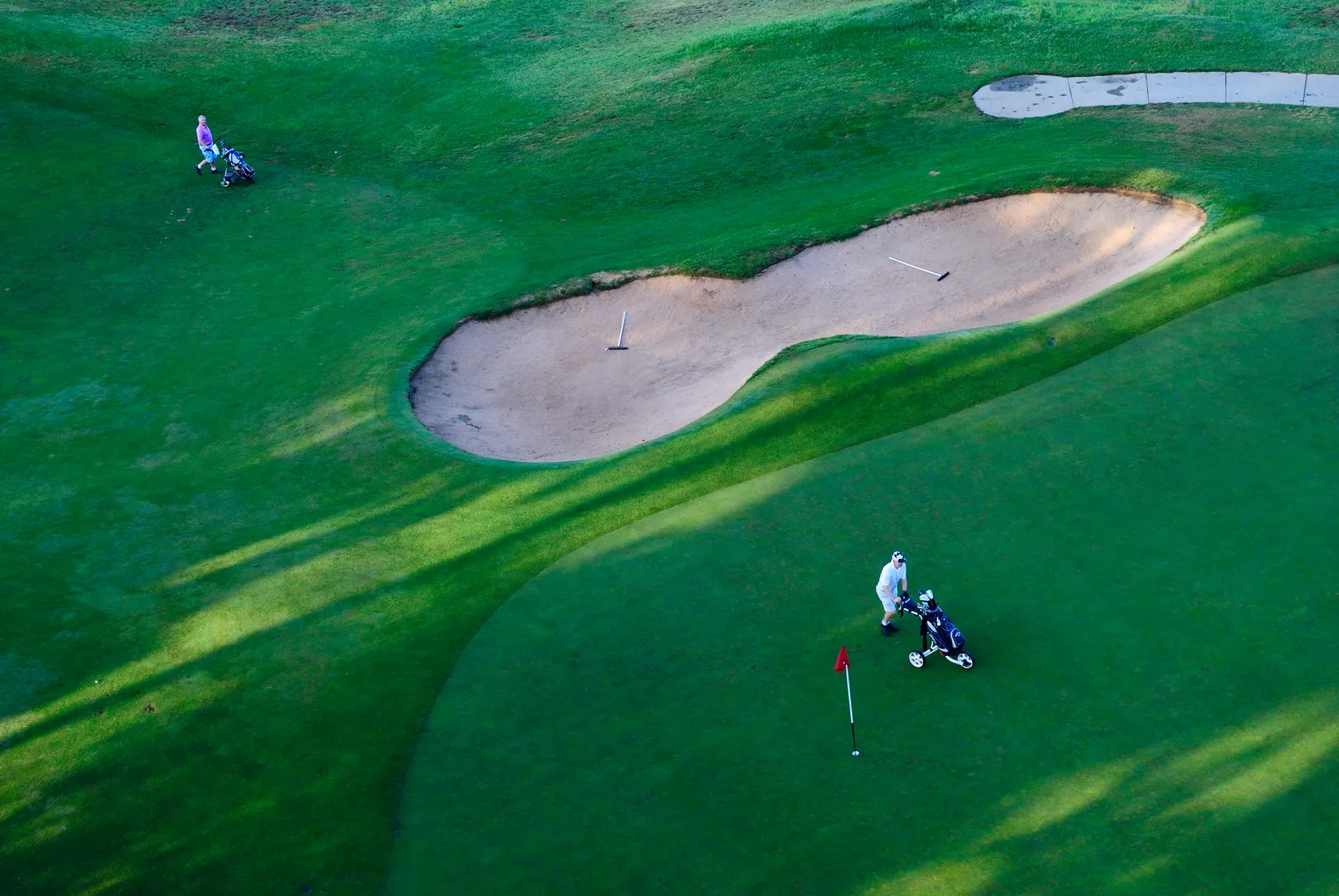 Green de golf vu d'en haut, avec un drapeau rouge, un bunker et un golfeur qui tire son chariot